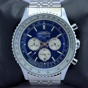 Könnte beinhalten: Eine Breitling Navitimer-Uhr aus Edelstahl mit blauem Zifferblatt und silbernen Hilfszifferblättern. Die Uhr verfügt über eine drehbare Lünette mit weißen Markierungen und ein Edelstahlarmband. Das Zifferblatt zeigt das Breitling-Logo und den Text "1884".