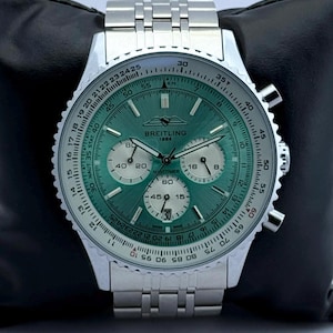 Könnte beinhalten: Eine silberne Breitling Navitimer Uhr mit grünem Zifferblatt. Die Uhr verfügt über mehrere Hilfszifferblätter, eine drehbare Lünette und ein Edelstahlarmband. Die Uhr wird auf einer schwarzen Oberfläche präsentiert.