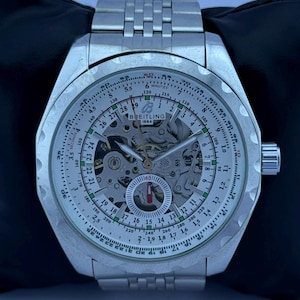 Op de afbeelding: Zilverkleurig Breitling 1884 horloge met een skeleton wijzerplaat, zichtbare tandwielen en een gedetailleerde lunette. Het horloge heeft een zilveren schakelband en een kroon aan de rechterkant. De wijzerplaat toont meerdere schalen en markeringen.