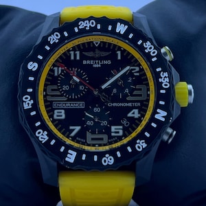Peut inclure: Une montre Breitling Endurance Chronometer avec un cadran noir, une lunette intérieure jaune et un bracelet en caoutchouc jaune vif. La lunette tournante noire présente des marquages de boussole blancs et le cadran affiche Breitling 1884, Endurance et Swiss Made.