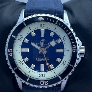 Pode incluir: Um relógio de pulso Breitling Superocean com mostrador azul, ponteiros e índices prateados e uma luneta preta rotativa. O relógio possui uma pulseira de borracha azul texturizada, uma janela de data e o texto Breitling 1884, Superocean, Chronometer 300m/1000ft, Swiss Made.