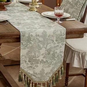 Puede incluir: Camino de mesa decorativo con un estampado toile de Jouy verde y blanco que representa un paisaje. El camino de mesa tiene un ribete dorado y borlas verdes. Se coloca sobre una mesa de madera.