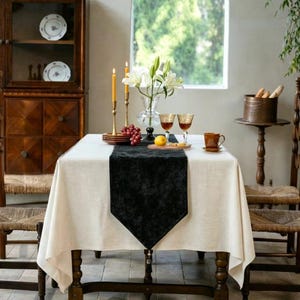 Pode incluir: Uma mesa de jantar retangular coberta com uma toalha de mesa de linho bege claro e um caminho de mesa central de veludo preto. A mesa está posta com dois castiçais de latão altos, um vaso de vidro com lírios brancos e dois copos âmbar.