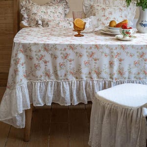May include: A floral tablecloth with a ruffled lace trim covers a wooden table. Matching floral pillows are on the chairs. A teacup, fruit, and a decorative glass are on the table. The tablecloth and pillows have a delicate pink and green floral pattern.