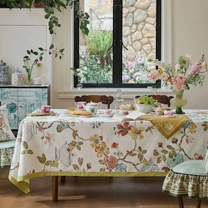 Pode incluir: Uma toalha de mesa branca com um padrão floral e botânico colorido com uma borda verde, sobre uma mesa de jantar posta com um bule, chávenas, uma taça de uvas e um vaso de flores, rodeada por cadeiras com capas de assento com folhos.