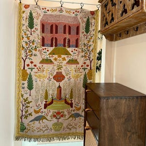 May include: A rectangular wall hanging featuring a cream-coloured fabric with a detailed embroidered scene of a pink house, cypress trees, yellow ducks, and various floral motifs. The piece has a yellow patterned border and a fringed bottom edge.