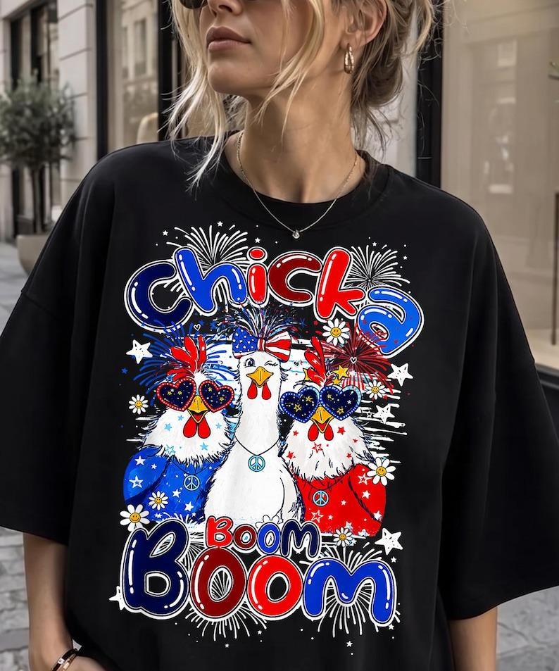 May include: A black short-sleeved t-shirt featuring a graphic print of three chickens wearing patriotic accessories. The text "Chicka Boom Boom" is displayed in blue and red. The design includes fireworks, stars, and flowers in red, white, and blue.