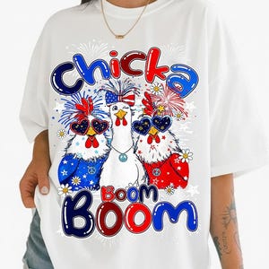 May include: A white short-sleeved t-shirt featuring a graphic of three chickens wearing patriotic accessories, including star-spangled sunglasses and headbands. The text "Chicka Boom Boom" is printed in blue and red bubble letters above and below.