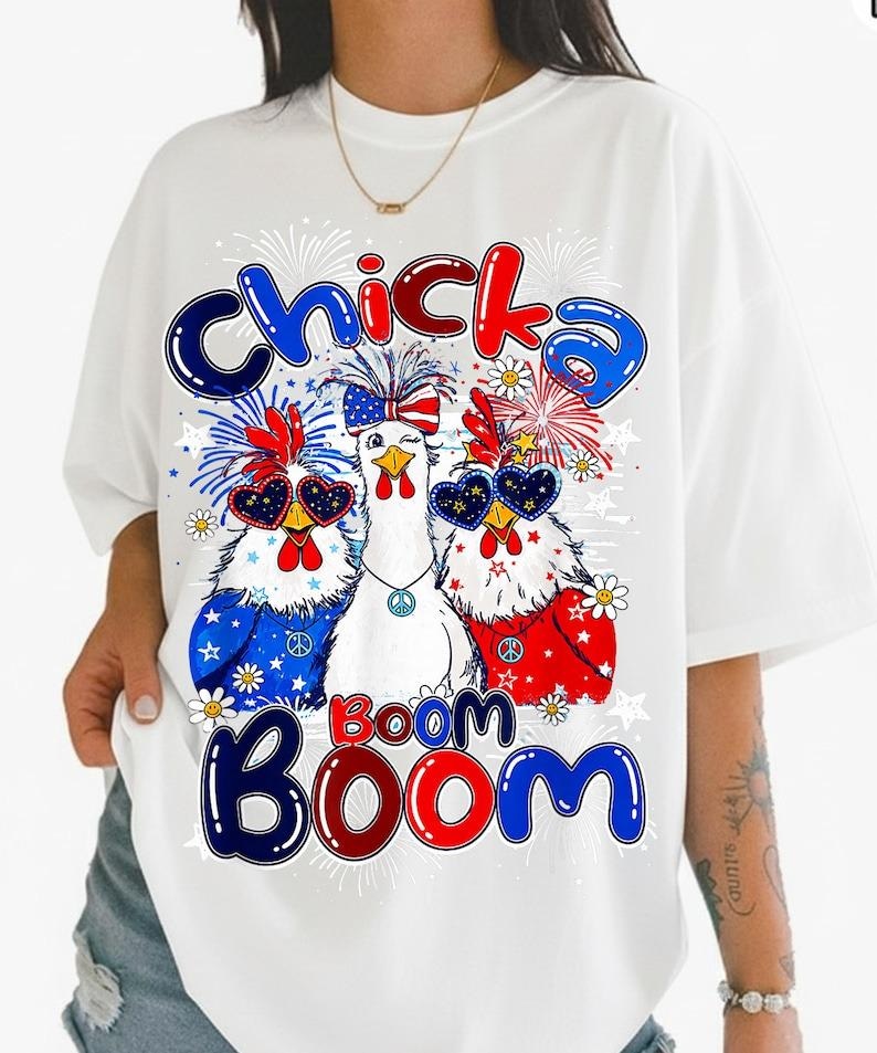 May include: A white short-sleeved t-shirt featuring a graphic of three chickens wearing patriotic accessories, including star-spangled sunglasses and headbands. The text "Chicka Boom Boom" is printed in blue and red bubble letters above and below.