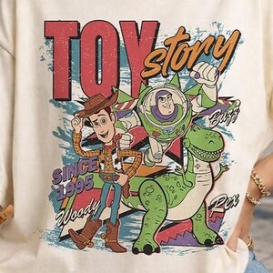 May include: A cream-coloured t-shirt featuring a graphic print of Woody, Buzz Lightyear, and Rex from Toy Story. The design includes the text "TOY story", "SINCE 1995", "Woody", "Buzz", and "Rex" in a distressed, colourful, and retro-inspired style.