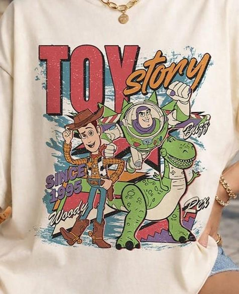 May include: A cream-coloured t-shirt featuring a graphic print of Woody, Buzz Lightyear, and Rex from Toy Story. The design includes the text "TOY story", "SINCE 1995", "Woody", "Buzz", and "Rex" in a distressed, colourful, and retro-inspired style.