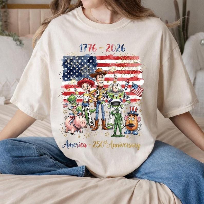 Op de afbeelding: Een cr&egrave;mekleurig t-shirt met een afbeelding van Toy Story-personages voor een Amerikaanse vlag met vuurwerk. De tekst luidt 1776-2026 en America-250th Anniversary. De personages zijn Woody, Buzz Lightyear, Jessie, Rex, Hamm, een groene soldaat en Mr. Potato Head.