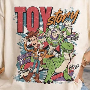 May include: A cream-coloured t-shirt featuring a graphic print of Woody, Buzz Lightyear, and Rex from Toy Story. The design includes the text "TOY story", "SINCE 1995", "Woody", "Buzz", and "Rex" in a distressed, colourful, retro-inspired font style.