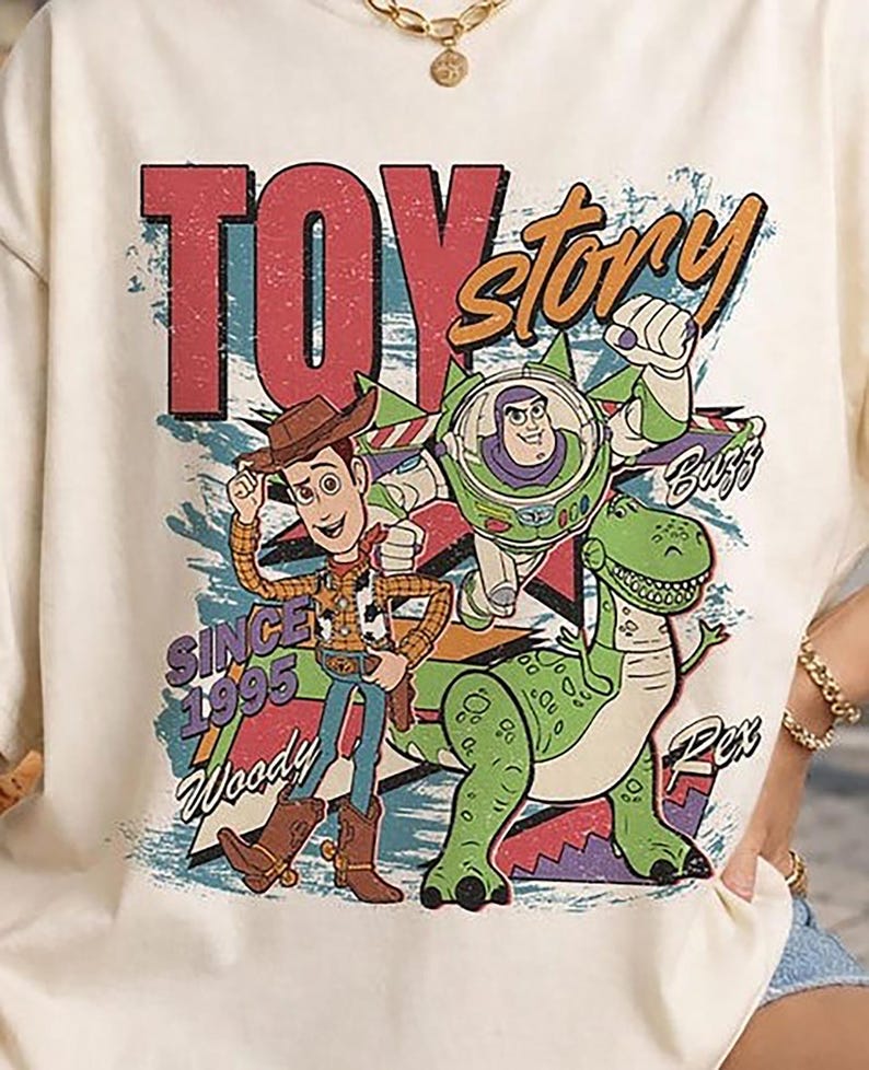 May include: A cream-coloured t-shirt featuring a graphic print of Woody, Buzz Lightyear, and Rex from Toy Story. The design includes the text "TOY story", "SINCE 1995", "Woody", "Buzz", and "Rex" in a distressed, colourful, retro-inspired font style.