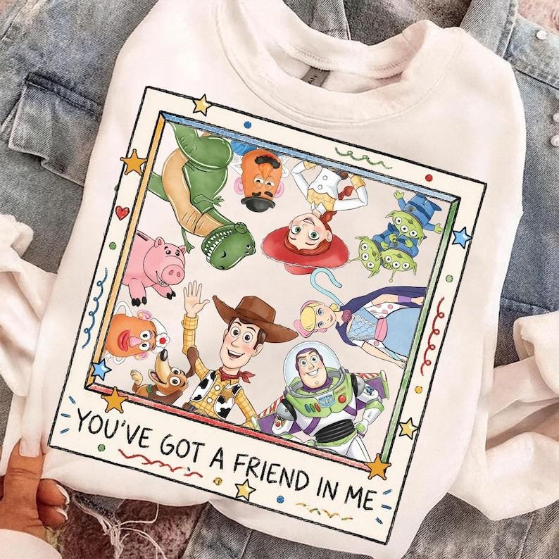 May include: A white crewneck sweatshirt featuring a square graphic print of various animated characters including a cowboy, a space ranger, a dinosaur, and a pig. The text "YOU'VE GOT A FRIEND IN ME" is printed in black at the bottom of the frame.