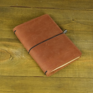 Puede incluir: Una funda rectangular para cuaderno de cuero en tono marrón medio, con una superficie lisa y mate de grano natural visible. Un cordón elástico negro fino envuelve horizontalmente el centro, asegurado por una pequeña cuenta metálica.