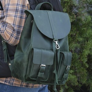 Può includere: Uno zaino in pelle verde scuro con chiusura a patta e due tasche frontali. Lo zaino ha una maniglia superiore e spallacci regolabili. L'hardware è color argento. La borsa è indossata da una persona che indossa una camicia a quadri e un gilet nero.