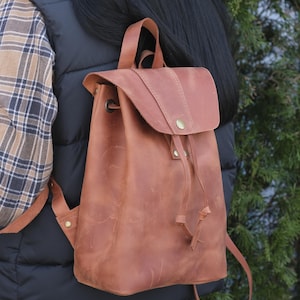 Può includere: Uno zaino in pelle marrone con chiusura a patta e coulisse. La borsa ha spallacci regolabili e una maniglia superiore. La pelle ha un aspetto invecchiato e l'hardware è color ottone. Lo zaino è indossato sopra un gilet nero e una camicia a quadri.