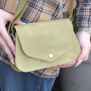以下が含まれることがあります： フラップ開閉とシルバーボタンが付いた、明るいオリーブグリーンのレザーショルダーバッグ。バッグにはステッチの縁取りと、お揃いの調節可能なストラップが付いています。バッグは、チェックのシャツとブルージーンズを着た人が持っています。