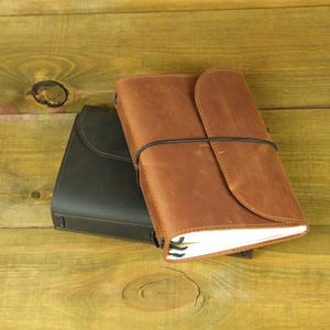 May include: Two leather journals, one in dark brown and one in light tan, each featuring a wrap-around cover design secured by a thin black elastic cord. The journals have visible white paper pages tucked inside the leather covers.
