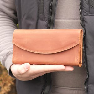 Könnte beinhalten: Eine hellbraune Ledergeldbörse mit geschwungenem Klappenverschluss. Die rechteckige Geldbörse ist handgenäht und hat eine natürliche, strukturierte Oberfläche. Die Geldbörse wird in der Hand einer Person gehalten.