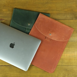 May include: A silver laptop with a black apple logo is positioned next to two flat rectangular sleeves. One sleeve is dark green and the other is tan, both featuring a rounded flap closure secured with a single silver metal snap button.