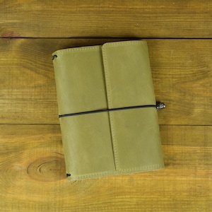 May include: A light olive green leather journal cover with a smooth, matte finish. The cover is secured by a thin black elastic cord that wraps horizontally around the center, featuring a small, dark metallic bead at the closure point.