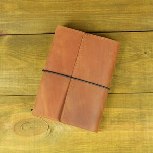 Puede incluir: Una funda rectangular para cuaderno hecha de cuero liso de color tostado. La funda presenta una solapa vertical en el lado derecho y está asegurada por una banda elástica negra delgada que se envuelve horizontalmente alrededor del centro.