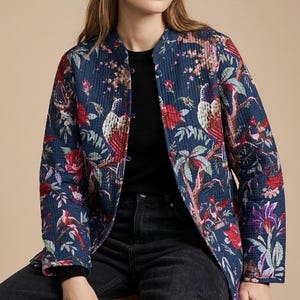 Könnte beinhalten: Eine marineblaue, gesteppte Jacke mit einem lebendigen botanischen Druck aus roten Blumen, violetten Blüten und Vögeln auf Ästen. Die Jacke hat einen Rundhalsausschnitt, lange Ärmel und eine Knopfleiste vorne mit passenden, stoffbezogenen Knöpfen.