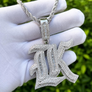 May include: A silver-toned pendant necklace featuring the letters "AK" encrusted with sparkling, clear gemstones. The pendant hangs from a silver rope chain. The background is a blurred green.