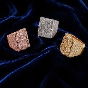 Puede incluir: Tres anillos cuadrados con diseños intrincados. Uno es de oro rosa, otro plateado y el tercero dorado. Cada anillo está cubierto de pequeñas piedras brillantes, creando una apariencia lujosa y llamativa. Los anillos se exhiben sobre una superficie de terciopelo azul oscuro.