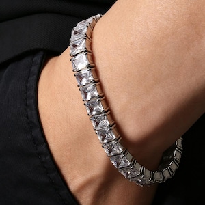 May include: A silver-toned tennis bracelet featuring a continuous row of square-cut, clear, faceted stones set in a metallic frame. The bracelet wraps around the wrist, showcasing a uniform design with reflective surfaces and a polished finish.