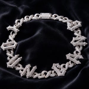 May include: A silver-toned, iced-out chain necklace with the word "NAVY" spelled out in ornate, diamond-studded letters. The necklace features a thick curb chain design and a secure clasp, set against a dark, textured background.
