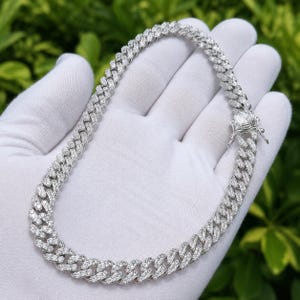May include: A silver-colored, iced-out chain necklace with a cuban link design. The necklace is encrusted with small, clear gemstones. The clasp is visible at the end of the necklace. The necklace is held in a gloved hand against a green background.
