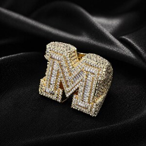 Op de afbeelding: Een goudkleurige ring in de vorm van de letter "M", bezet met kleine, sprankelende edelstenen. De ring is geplaatst tegen een zwarte achtergrond, wat het ingewikkelde ontwerp en de luxueuze uitstraling benadrukt. De ring is een statement-sieraad.