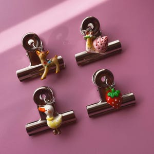 May include: Four silver metal binder clips, each featuring a small dangling charm. The charms include a brown deer with floral details, a pink snail, a yellow duck with white polka dots, and a red strawberry with green leaves.
