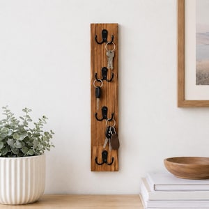Puede incluir: Un soporte para llaves de pared rectangular y vertical de madera con cuatro ganchos dobles de metal negro mate. Se aprecia la veta natural de la madera y varios llaveros de metal con llaves cuelgan de los ganchos en línea.