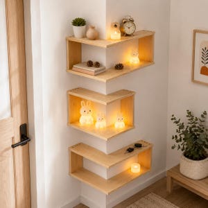 Puede incluir: Tres estantes de esquina de madera clara montados en una pared blanca. El estante superior contiene una pequeña planta en maceta, un jarrón, una vela y un reloj. El estante central presenta tres lámparas brillantes con forma de conejo.