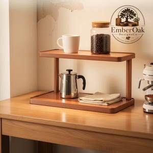 Puede incluir: Una estantería de madera de dos niveles con una configuración de café. La balda superior contiene una taza blanca y un tarro de cristal lleno de granos de café. La balda inferior tiene una cafetera de acero inoxidable y un paño beige doblado.