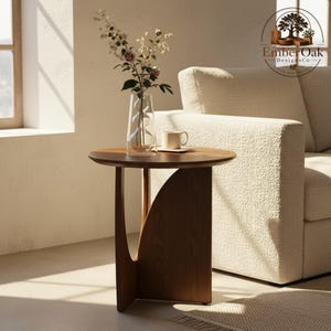 May include: A round, dark brown wooden side table with a unique curved leg design. The table has a vase with flowers and a coffee cup on top. The Ember Oak DesignsCo logo is in the upper right corner.