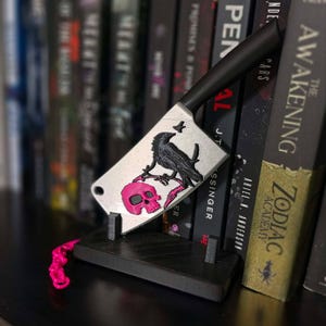 Peut inclure: Un objet décoratif en forme de couperet avec un manche noir et une lame argentée ornée d'un corbeau noir perché sur une branche au-dessus d'un crâne rose vif. Le couperet repose sur un support rectangulaire noir avec une petite chaîne rose.