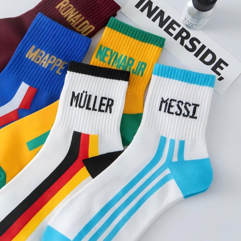Peut inclure: Une collection de chaussettes de sport de diff&eacute;rentes couleurs, chacune portant le nom d'un joueur de football en lettres grasses. Les chaussettes sont blanches, bleues, jaunes et bordeaux, avec des accents tels que des rayures et des blocs de couleur. Le mot "INNERSIDE" est visible en arri&egrave;re-plan.