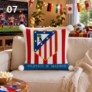 K&ouml;nnte beinhalten: Ein quadratisches Zierkissen mit dem Logo des Fu&szlig;ballvereins Atletico de Madrid, roten und wei&szlig;en vertikalen Streifen, einem blauen Rand und wei&szlig;en Bommeln an jeder Ecke. Der Text ATLETICO DE MADRID ist unten in Wei&szlig; aufgedruckt.