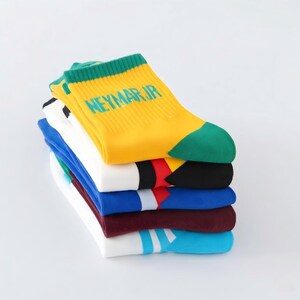 Peut inclure: Une pile de chaussettes color&eacute;es. La chaussette du dessus est jaune avec des accents verts et le texte "NEYMAR JR". D'autres chaussettes sont blanches, bleues et bordeaux, avec des rayures et des blocs de couleur. Les chaussettes sont empil&eacute;es sur une surface blanche.