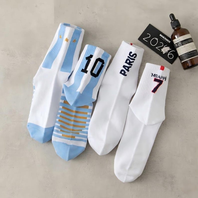 Puede incluir: Cuatro pares de calcetines deportivos blancos. Dos pares presentan rayas azul claro y el n&uacute;mero "10" o estrellas doradas. Los otros dos pares tienen la palabra "PARIS" y el n&uacute;mero "7" con el nombre "MBAPPE" en negro.