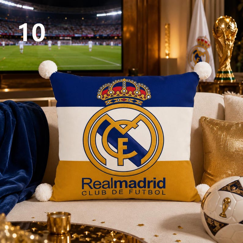 K&ouml;nnte beinhalten: Ein quadratisches Dekokissen mit dem Logo des Real Madrid Club de Futbol, einer Krone und dem Schriftzug Realmadrid Club de Futbol. Das Kissen hat ein horizontales dreifarbiges Design in Blau, Wei&szlig; und Gold mit wei&szlig;en Bommeln an den Ecken.