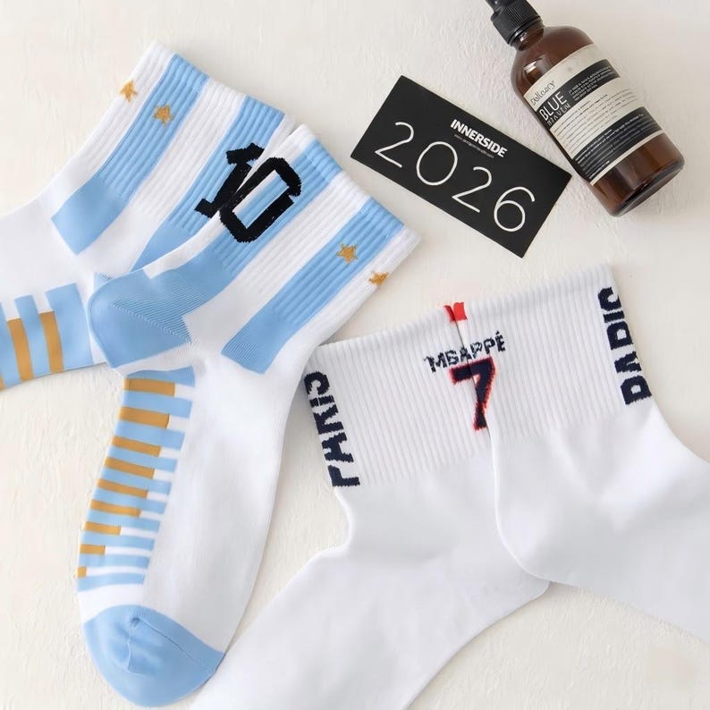 Puede incluir: Calcetines deportivos blancos con rayas azules y doradas y el n&uacute;mero 10. Otro par de calcetines blancos presenta el nombre "Mbapp&eacute;" y el n&uacute;mero 7 en rojo. Una tarjeta negra dice "2026" e "Innerside".