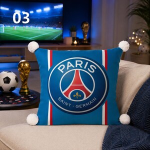 K&ouml;nnte beinhalten: Ein quadratisches blaues Dekokissen mit dem Logo des Fu&szlig;ballvereins Paris Saint-Germain in der Mitte, flankiert von vertikalen roten und wei&szlig;en Streifen. Das Kissen hat an jeder der vier Ecken einen wei&szlig;en Bommel, was f&uuml;r eine dekorative Textur sorgt.