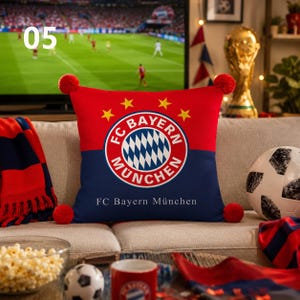 K&ouml;nnte beinhalten: Ein quadratisches Zierkissen mit dem FC Bayern M&uuml;nchen Logo in Rot und Marineblau. Das Design zeigt vier gelbe Sterne &uuml;ber dem runden Wappen und den Schriftzug FC Bayern M&uuml;nchen in Wei&szlig; am unteren Rand, erg&auml;nzt durch rote Bommeln an den Ecken.