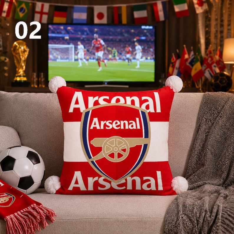 K&ouml;nnte beinhalten: Ein quadratisches rot-wei&szlig;es Zierkissen mit dem Arsenal-Fu&szlig;ballclub-Logo in der Mitte und dem Wort Arsenal, das &uuml;ber und unter dem Wappen aufgedruckt ist. Das Kissen hat vier wei&szlig;e Bommeln an den Ecken und eine weiche Stofftextur.